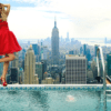 A woman in red dress standing on edge of pool with city skyline.